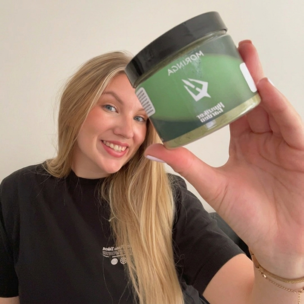 Mosolygó szőke nő fekete pólóban a HunzikerGarden Moringa zöld por tégelyét tartja az arca mellett.